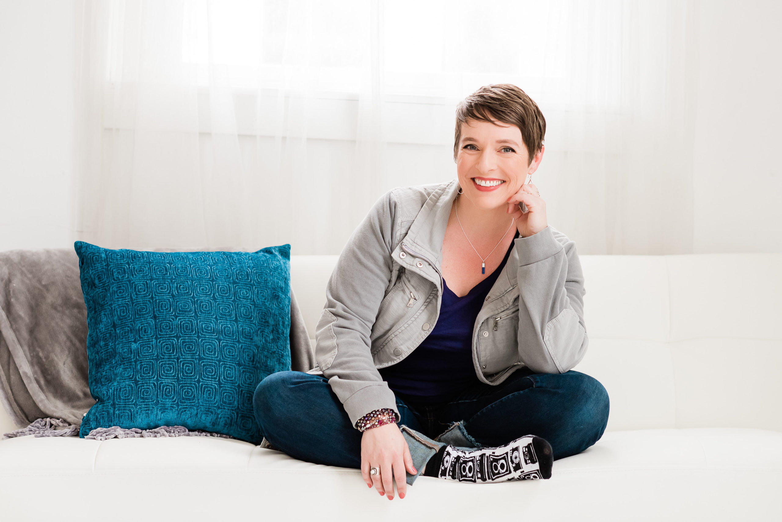 Smiling woman sitting cross legged on a white couch with blue pillow.